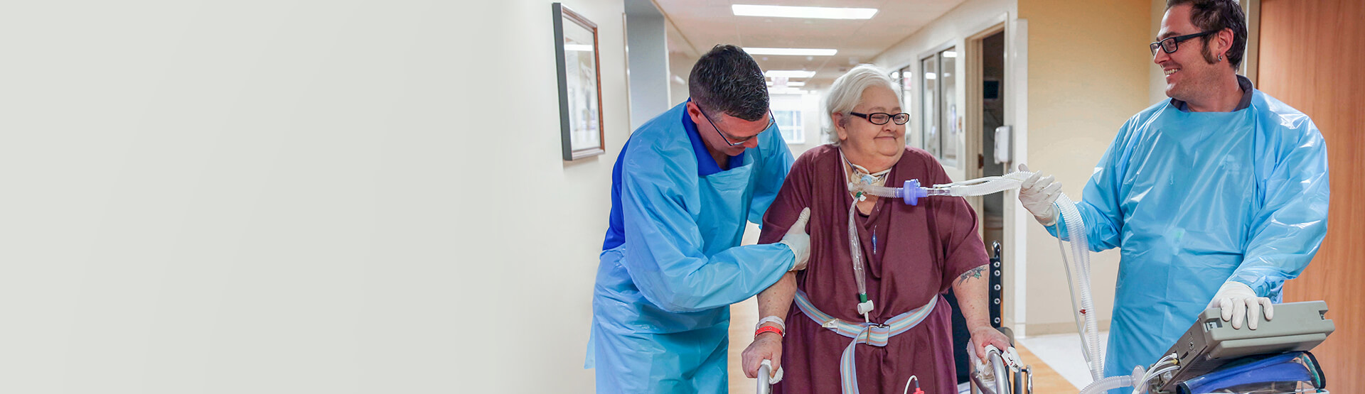 Older female patient standing with a walker while a respiratory therapist holds her ventilator airway tube.