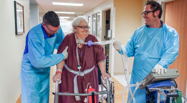 Older female patient standing with a walker while a respiratory therapist holds her ventilator airway tube.