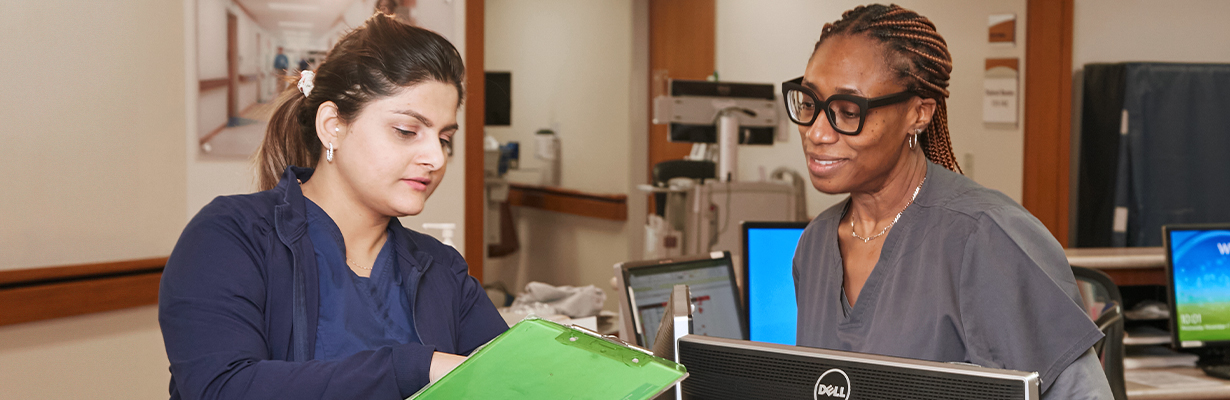 Two nurses gather at the nurses station looking at a clipboard discussing patient care