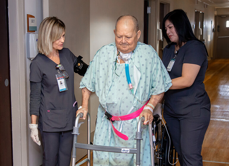 A male patient wearing a hospital gown and on a ventilator uses the support of a walker and his nurses to walk down the hall.