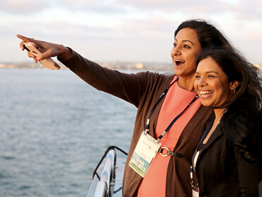 Two female Select Medical employees enjoy the view while on a dinner cruise during a conference. Both smiling at the horizon, one pointing while holding her phone.