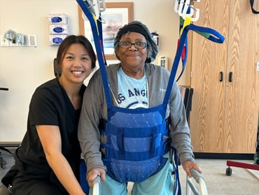 Carol Roseboro utilizes supportive equipment and her walker to walk. She smiles at the camera with her nurse.