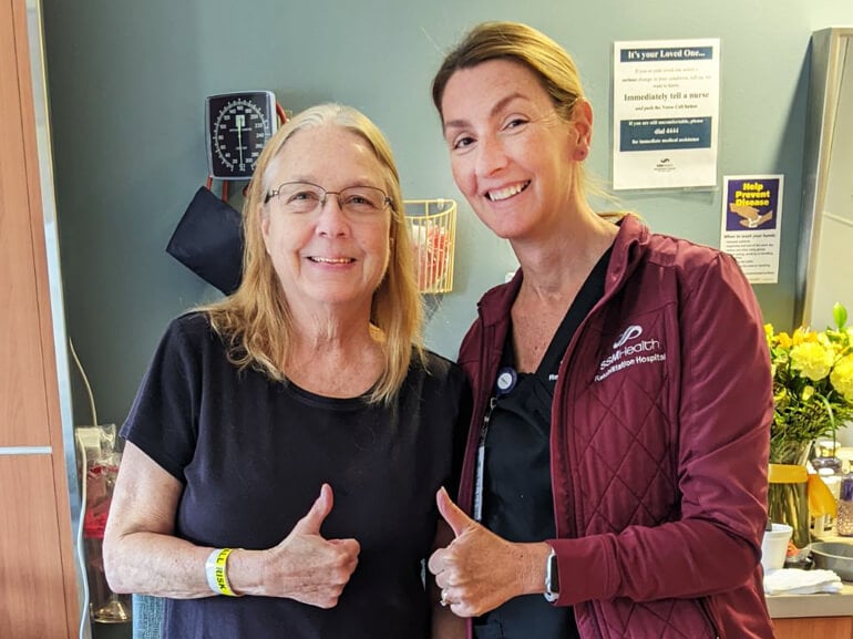 Cindy and her nurse stand in her hospital room, both smiling and giving the camera a thumbs up.