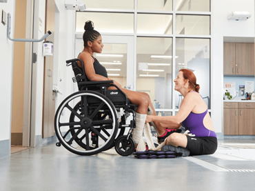 Kenya sits in her wheelchair by a therapy pool while a therapist helps to wrap her legs. The two smile at eachother.