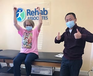 Judy sits on a medical table in front of a Rehab Associates sign next to her physical therapist. She cheers with her arms up and her physical therapist gives a thumbs up.