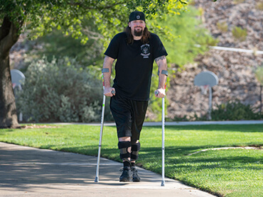 Logan uses crutches to walk down a path at a park. He smiles as he walks towards the camera.