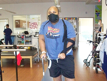 Jean, wearing a sling on his left arm, holds a cane in his right hand that he uses to help him walk.